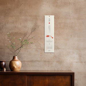 A vertical Japanese calendar poster with a white background, black kanji text, and a red koi fish illustration, displayed on a textured beige wall above a dark wooden cabinet with a ceramic vase.