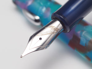 Close-up of a fountain pen nib and upper barrel showing a polished silver nib with gold script engraving, attached to a translucent blue and purple marbled resin cap on a light gray background.