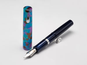 A fountain pen and its cap displayed on a light gray background. The pen body is dark navy blue with silver metal accents, and the cap features an iridescent marbled pattern with blue, purple, and green tones.