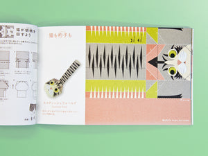 An open Japanese craft book laid flat on a mint green background, showing printed folding instructions on one page and a colorful assembled paper cat toy on the other.