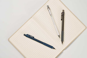 An open lined notebook on a white background with three pens placed on top: a dark blue pen, a white pen with silver accents, and a dark gray/black pen.