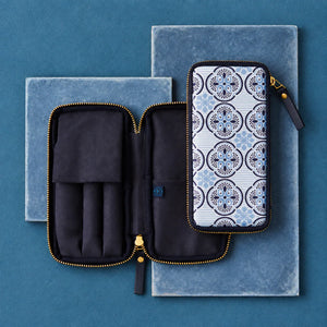A zippered travel or organizer pouch shown open and closed on top of blue velvet boxes. The pouch interior is black with gold-tone zippers, and the exterior has a white, blue, and gray geometric medallion pattern.