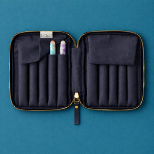 An open zippered jewelry organizer case in dark navy fabric with gold-tone zipper trim, laid flat on a teal background. The case has multiple vertical slots, a flap pocket, and two small gemstone or crystal pieces stored inside.