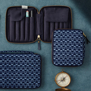 A set of navy blue zippered travel or makeup cases arranged on a textured teal background, including an open multi-compartment case, a smaller matching zip case, and a flat rectangular pouch with a geometric pattern. A vintage-style compass is placed near the bottom right.