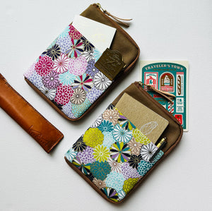 Two small zip-around card holders with brown leather trim and a colorful floral patterned fabric front, shown on a light background with inserted cards and a matching leather strap.