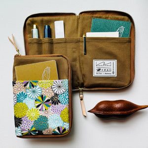 An open brown canvas zippered organizer pouch with multiple interior pockets, shown alongside a matching closed pouch featuring a colorful floral print. A small wooden leaf-shaped object sits to the right on a light neutral background.