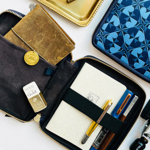 An open black zippered travel organizer wallet with gold-tone zippers, shown in a styled flat-lay on a white background. Inside are a cream notebook, a gold pen, a blue pen, and a gold coin.