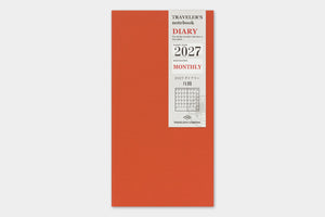 A vertical orange-red traveler's notebook with a white paper label on the front showing '2027 Monthly' and 'Traveller's Notebook' text, photographed against a light gray background.