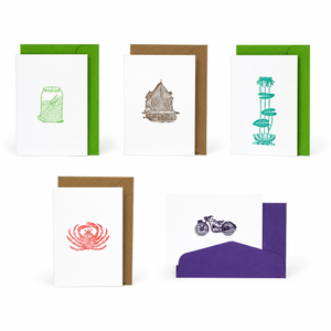 Five greeting cards with envelopes arranged on a white background. The cards feature minimalist illustrations in green, brown, teal, red, and purple, including a jar, a house, water lilies, a crab, and a motorcycle.