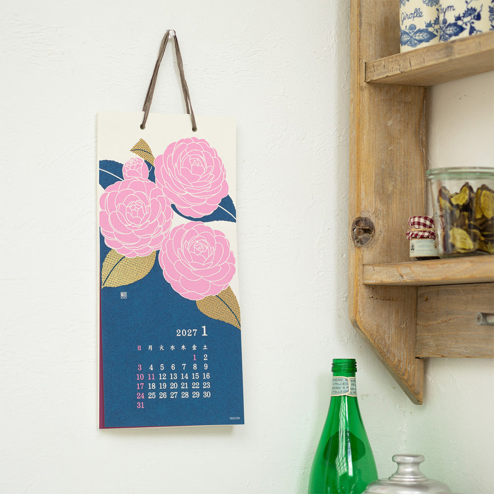 A vertical hanging calendar page with a blue background, pink flowers, and a small 2027 January calendar grid, displayed on a white wall next to a wooden shelf and green bottles.