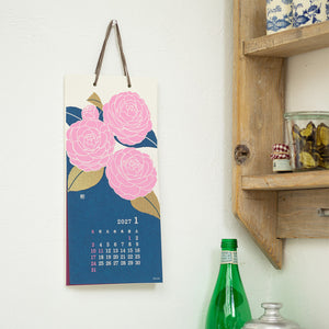 A vertical hanging calendar page with a blue background, pink flowers, and a small 2027 January calendar grid, displayed on a white wall next to a wooden shelf and green bottles.