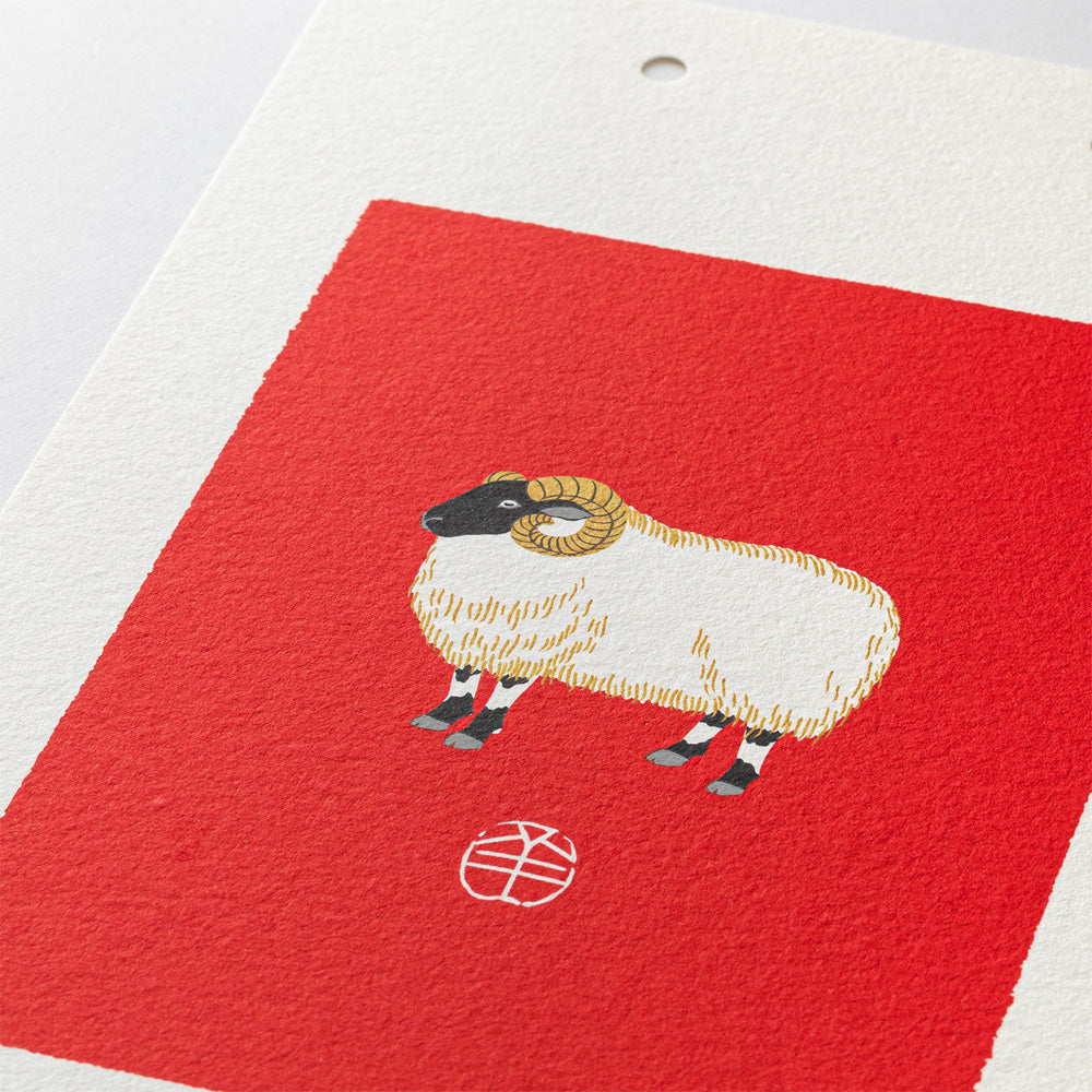A close-up of a textured white paper card featuring a red square print with an illustration of a white sheep with a curled golden-yellow horn and a small white circular emblem below it.