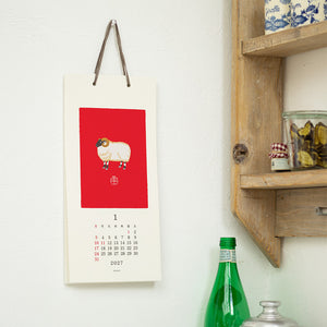 A vertical wall calendar hanging on a white wall, featuring a red square with a sheep illustration and the date grid for 2017. Part of a wooden shelf with jars and a green bottle is visible on the right.
