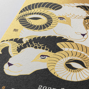 Close-up of a Japanese-style art print featuring two ram heads with large curled horns in gold, black, and off-white, with part of a dark textured surface and small white numerals visible below.