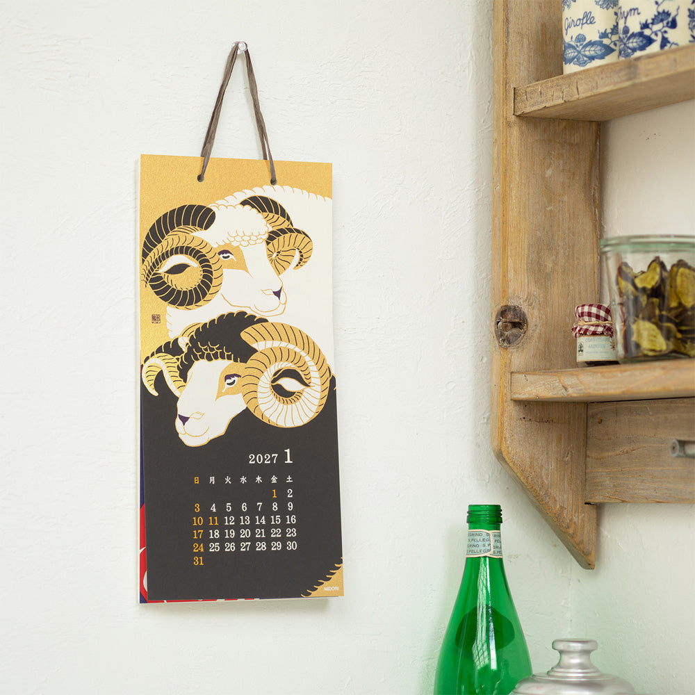 A vertical 2027 January wall calendar featuring two stylized ram illustrations in gold, black, and cream, hanging on a white wall next to a wooden shelf with decorative items.