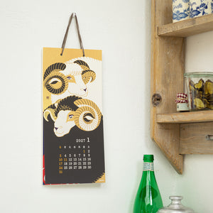 A vertical 2027 January wall calendar featuring two stylized ram illustrations in gold, black, and cream, hanging on a white wall next to a wooden shelf with decorative items.