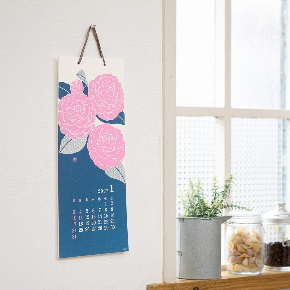 A vertical wall calendar hanging on a white wall, featuring large pink flowers on a blue background with a small calendar grid for January. The calendar is styled in a bright room next to a window, with small potted plants and jars on a wooden shelf.