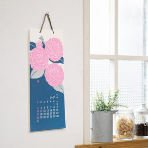 A vertical wall calendar hanging on a white wall, featuring large pink flowers on a blue background with a small calendar grid for January. The calendar is styled in a bright room next to a window, with small potted plants and jars on a wooden shelf.