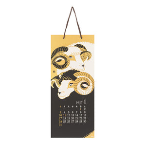 A hanging wall calendar page featuring two stylized ram illustrations in gold, cream, and black, with a small 2027 January calendar grid at the bottom and a dark brown hanging loop at the top.