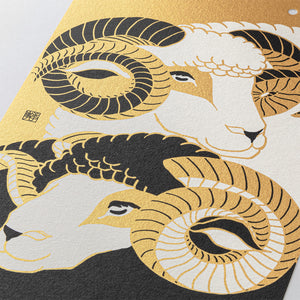 Close-up of a decorative art print featuring stylized ram heads and curled horns in gold, black, and off-white on a textured surface.