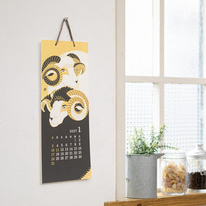 A vertical wall calendar hanging on a white wall, featuring two stylized ram illustrations in yellow, black, and white, with the 2021 January calendar dates printed near the bottom. The calendar is shown in a bright room next to a window, with small potted plants and jars on a wooden surface below.