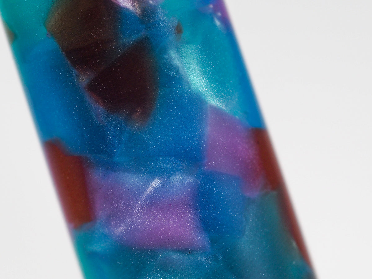A close-up of a cylindrical product, likely a pen or tube, featuring a marbled pattern of metallic blue, purple, and red with a glittery finish, set against a plain white background.