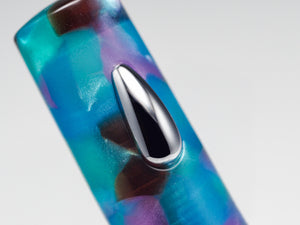 Close-up of a translucent acrylic or glass-like cylindrical product with swirling blue, teal, purple, and brown marbled colors, featuring a prominent polished silver teardrop-shaped inlay or accent on a light gray background.