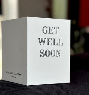A white square greeting card or box with the words 'GET WELL SOON' printed in a dark gray serif font on the front, photographed on a dark fabric surface with a softly blurred indoor background.