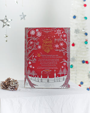 A red Advent calendar box with silver winter tree illustrations and gold lettering, displayed on a white draped surface with a pinecone and softly blurred holiday decorations in the background.
