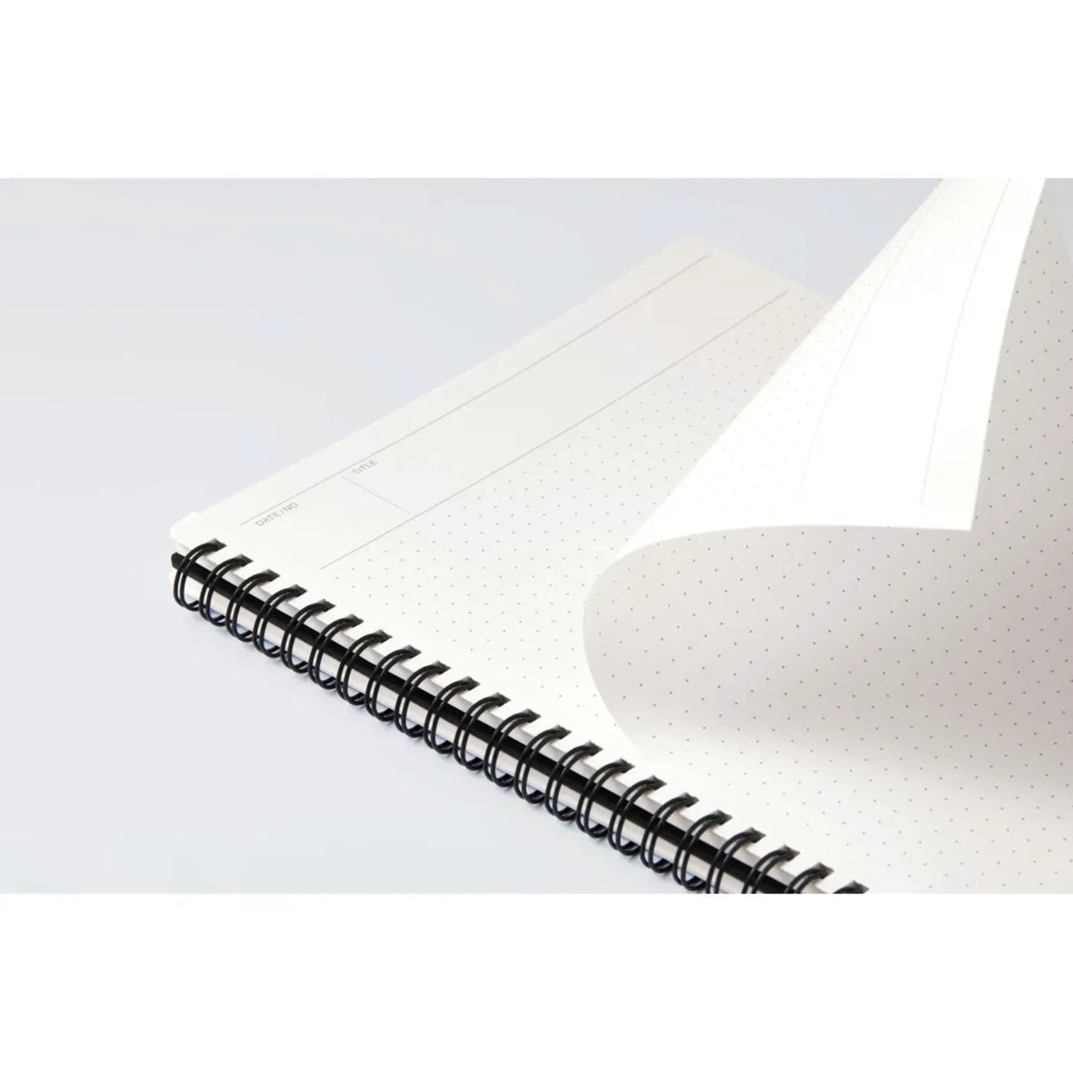 A close-up of an open spiral-bound notebook with a black wire coil and white dotted page, showing a blank lined page being turned over on a light gray background.