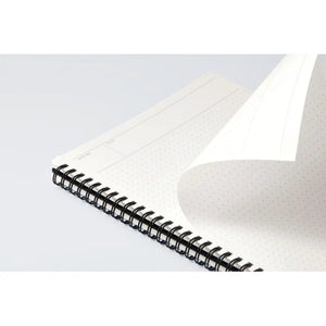 A close-up of an open spiral-bound notebook with a black wire coil and white dotted page, showing a blank lined page being turned over on a light gray background.