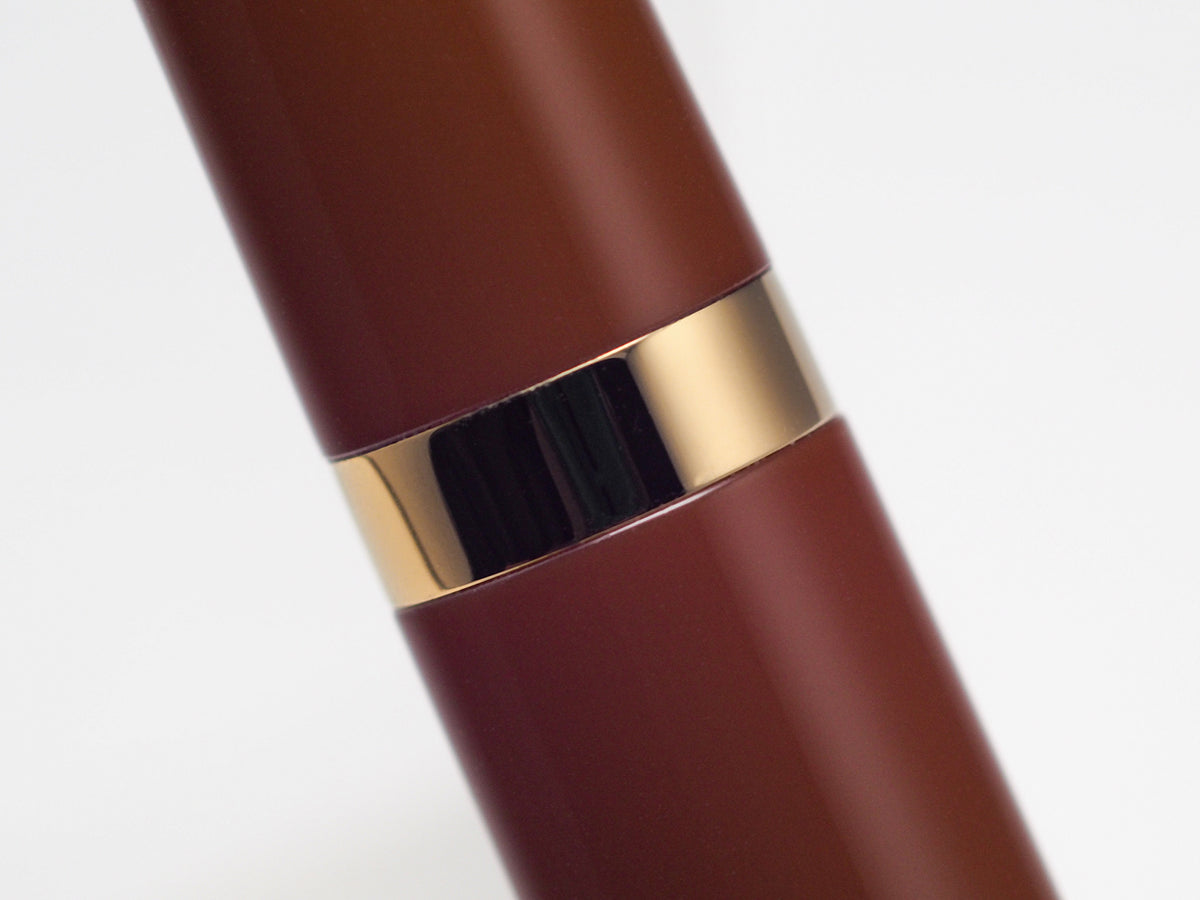 Close-up of a dark reddish-brown cylindrical product, likely a pen or tube, with a shiny gold metallic band around the middle, set against a plain white background.