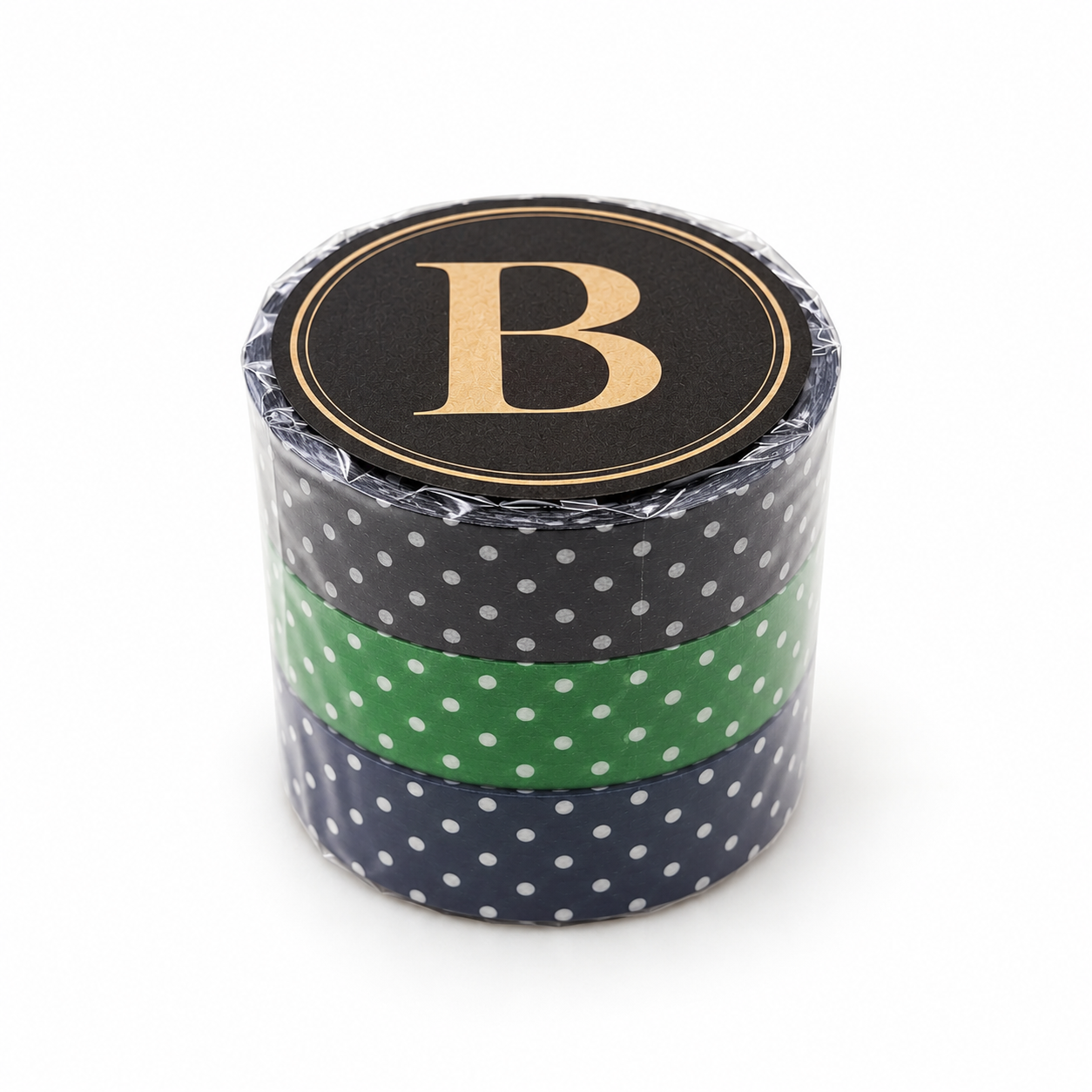 A stack of three decorative washi tapes with white polka dots on a navy blue base and a solid green middle tape, wrapped in clear plastic with a black lid featuring a gold letter 'B'.
