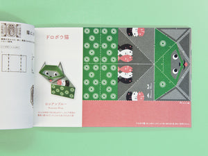An open Japanese origami booklet or craft book laid flat on a mint green background, showing illustrated pages with folding instructions and a completed green cat origami model.