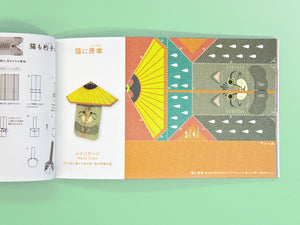 An open Japanese craft book or instruction booklet laid flat on a mint green background. The right page shows a colorful illustrated patchwork design with a cat face and a yellow umbrella motif, while the left page contains sewing or construction diagrams.