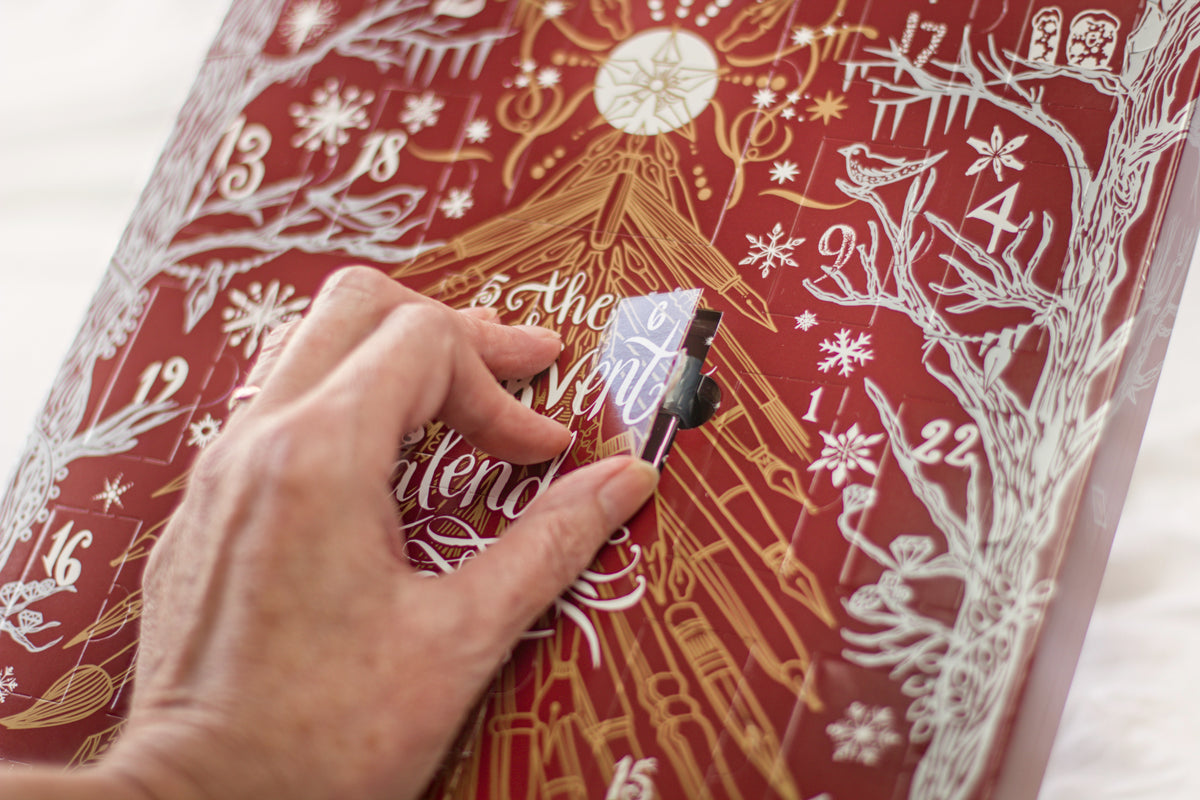 A close-up of a hand peeling back a layer of a red advent calendar with gold and white illustrations and text.