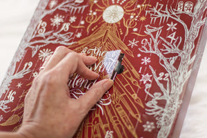 A close-up of a hand peeling back a layer of a red advent calendar with gold and white illustrations and text.
