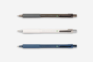 Three retractable gel pens arranged horizontally on a light gray background: a dark gray pen with a brown barrel, a white pen with a silver clip, and a slate blue pen with a dark blue barrel.