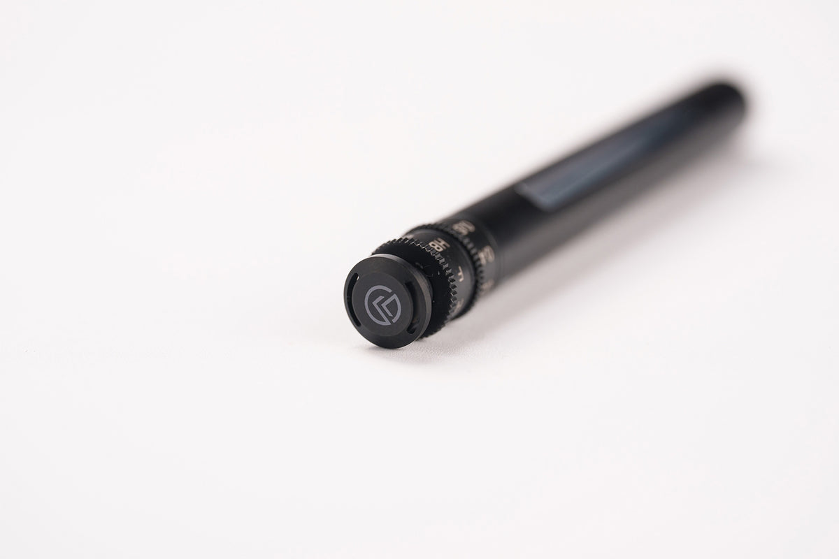 A close-up of a black cylindrical camera lens focusing ring with a textured adjustment knob and a circular logo, set against a plain white background.