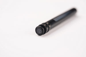 A close-up of a black cylindrical camera lens focusing ring with a textured adjustment knob and a circular logo, set against a plain white background.