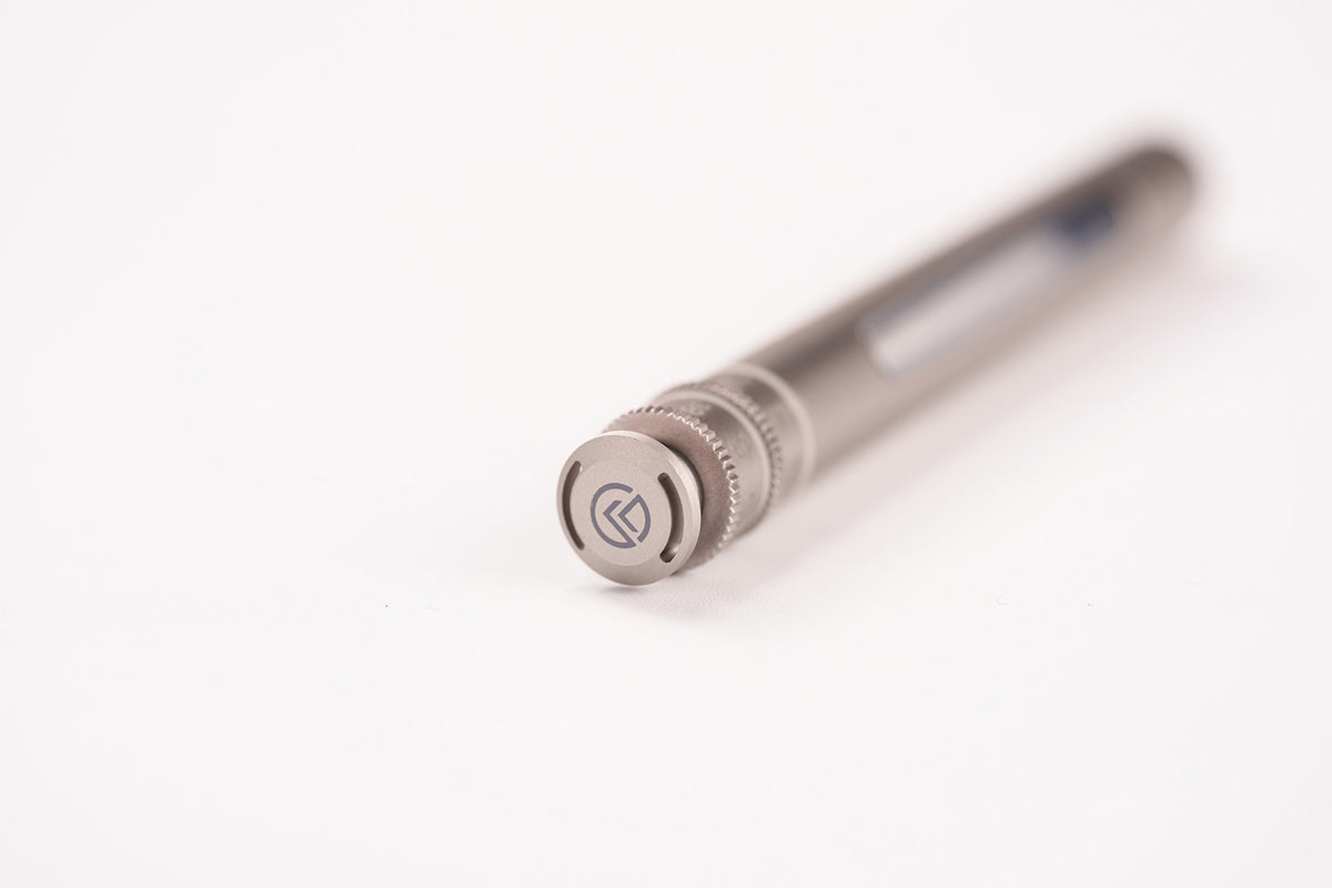 A close-up of a metallic silver pen with a textured knurled cap and a circular logo emblem, resting on a plain white surface with a shallow depth of field.