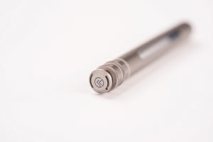 A close-up of a metallic silver pen with a textured knurled cap and a circular logo emblem, resting on a plain white surface with a shallow depth of field.