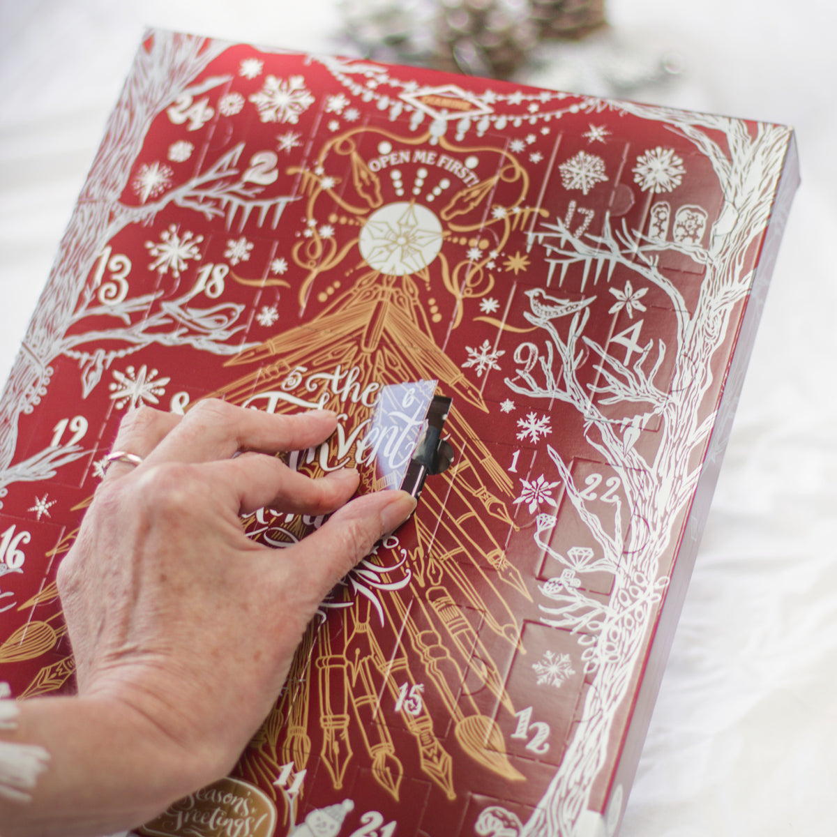 A close-up of a hand opening a red Christmas advent calendar with gold and white illustrations and numbered windows.