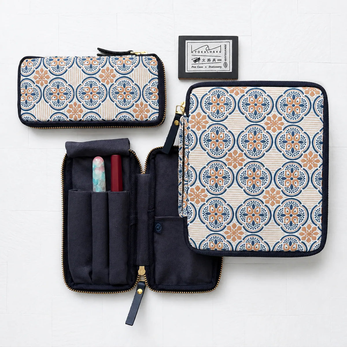 Three views of a zippered fabric organizer case with a blue and orange geometric medallion pattern on a cream background, shown closed and open with interior pockets and accessories, plus a small brand card on a white background.