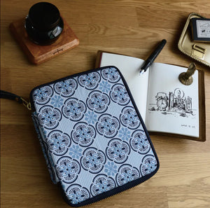A zippered pouch with a blue and white geometric medallion pattern, styled on a wooden desk next to an open notebook, a black pen, and other stationery items.