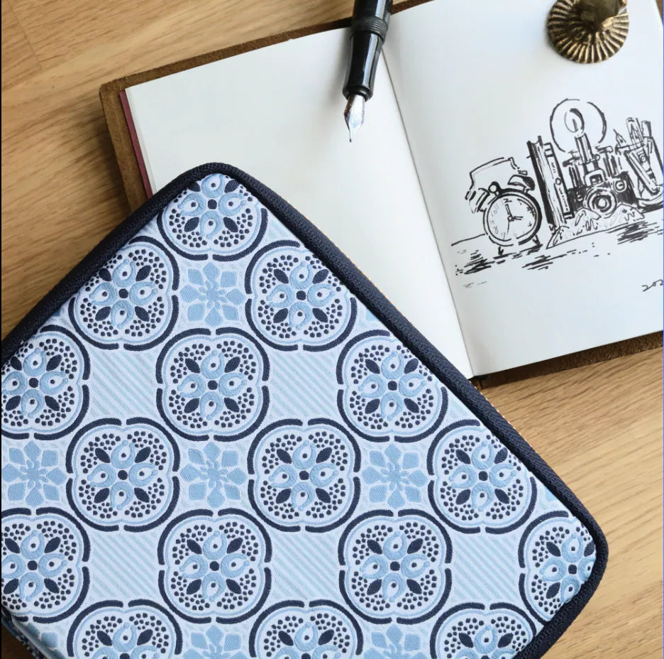 A square fabric item with a repeating blue and gray floral medallion pattern and black trim, laid on a wooden surface next to an open notebook with a pen.