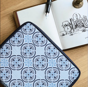 A square fabric item with a repeating blue and gray floral medallion pattern and black trim, laid on a wooden surface next to an open notebook with a pen.