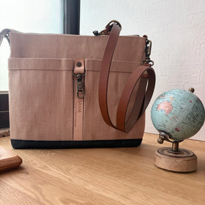 A canvas shoulder bag in a dusty pink-beige color with brown leather straps and a black base, displayed on a wooden table next to a small globe.