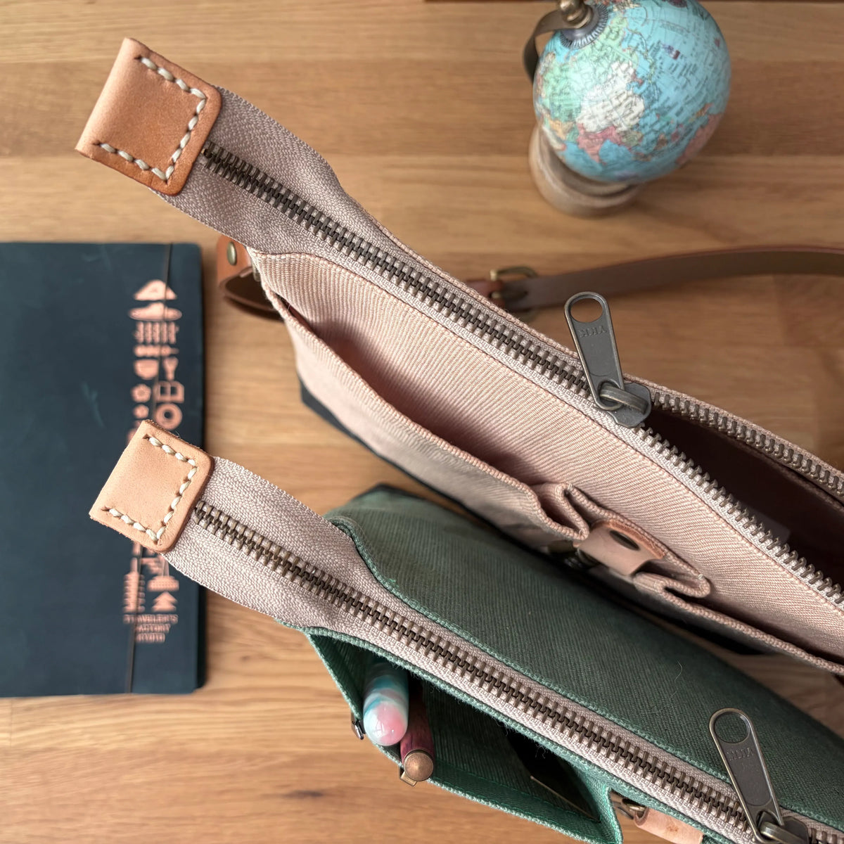 Two canvas zippered pouches laid on a wooden surface, one in beige and one in dark green, with a notebook and a small globe in the background.