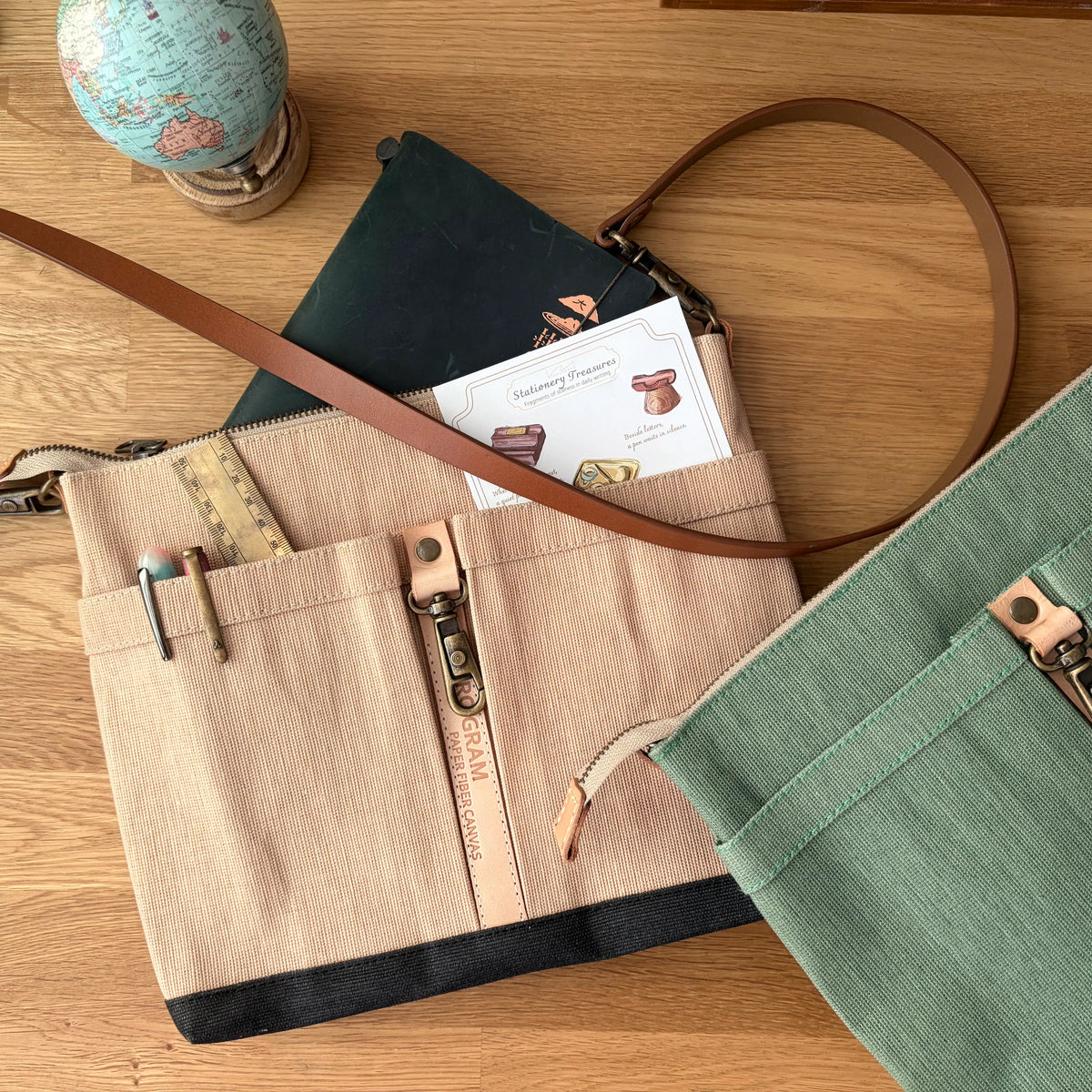 A beige canvas crossbody bag with brown leather trim and a long strap, styled on a wooden tabletop with a green notebook, a globe, and another green bag partially visible.